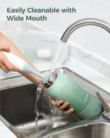 Momcozy portable breast milk warmer, mint green, being cleaned under a faucet. Features wide mouth design for easy cleaning.