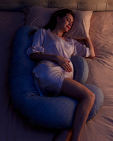 Pregnant woman side-sleeping on gray Momcozy F-shaped pregnancy pillow for full body support and comfortable sleep.
