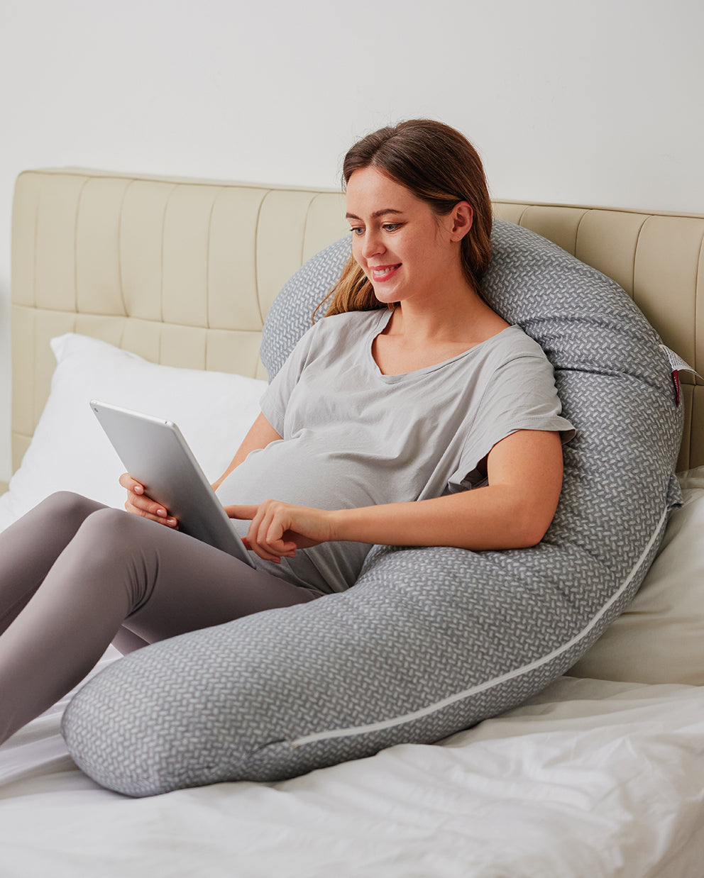 Pregnant woman using tablet, side-sleeping on gray Momcozy F-shaped pregnancy pillow for comfort and support.