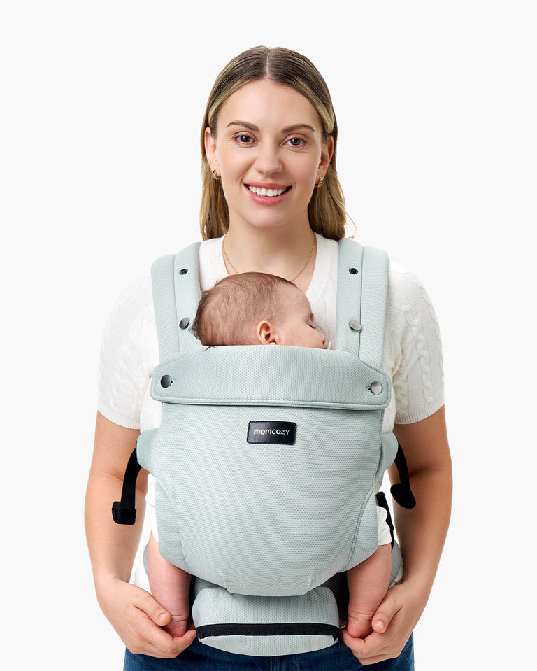 Momcozy baby carrier in light blue, supporting a sleeping baby, worn by a smiling parent, featuring the Momcozy logo.